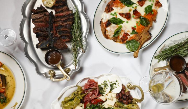 Food like steak and pizza on platters atop a white tablecloth.